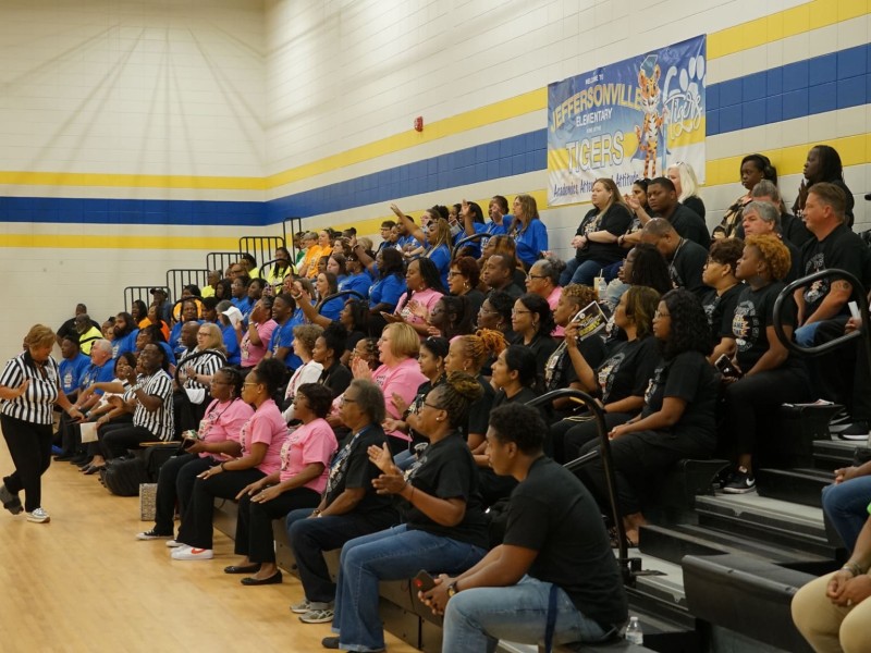 People wearing green, black, pink and blue sit in gym stands in sections cheering. A few people wearing black and white stripped referee shirts stand in front of the stands. Behind the stands is a banner that says "Welcome to Jeffersonville Elementary."