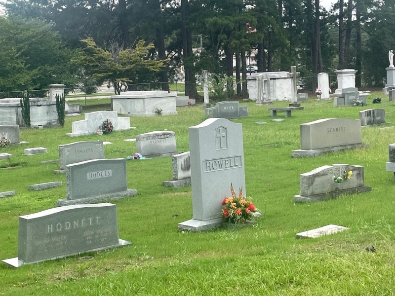 Many gravestones in green grass in a cemetery. They vary in size, and some have flowers and small American flags in front of them.